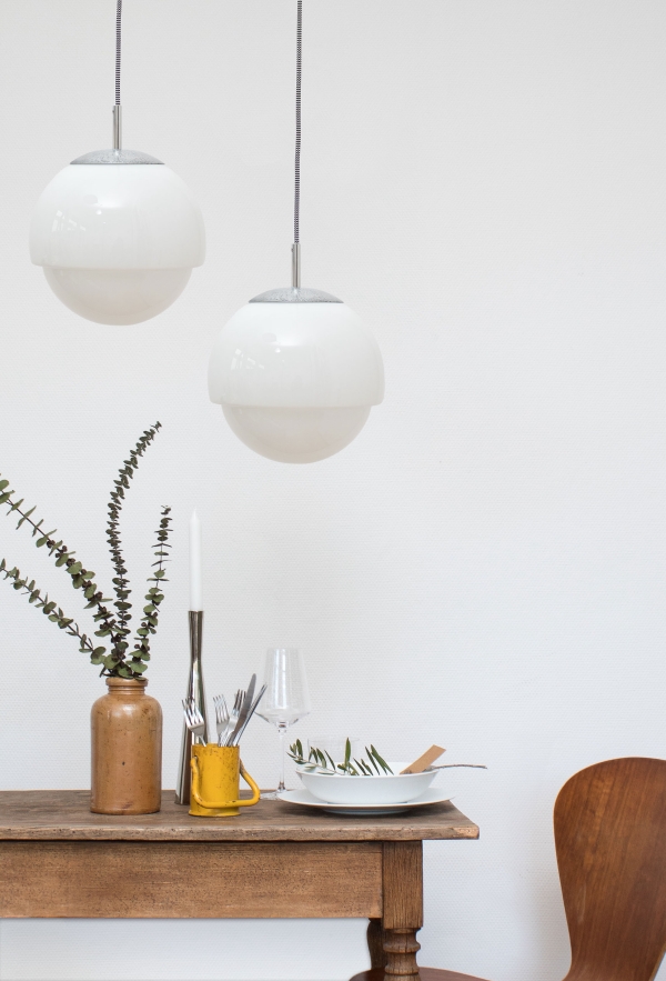 Ein kleiner Holztisch, gedeckt mit einem weißen Teller, einem Weinglas, einem silbernen Kerzenständer und einer braunen Vase mit Eukalyptuszweigen darin. Rechts vom Tisch sieht man noch einen kleinen teil eines Stuhls. Über dem Tisch hängen zwei elegante