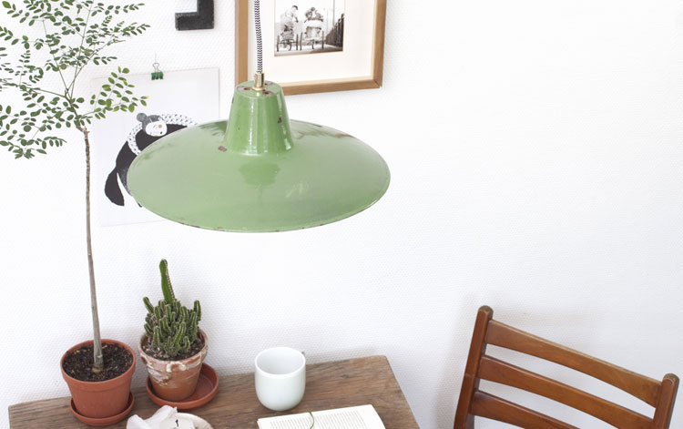 Im Bildausschnitt ist ein Teil eines altes Holztisches zu sehen, darauf stehen zwei Grünpflanzen und eine Kaffeetasse. Über dem Tisch hängt eine Lampe aus grünem Emaille