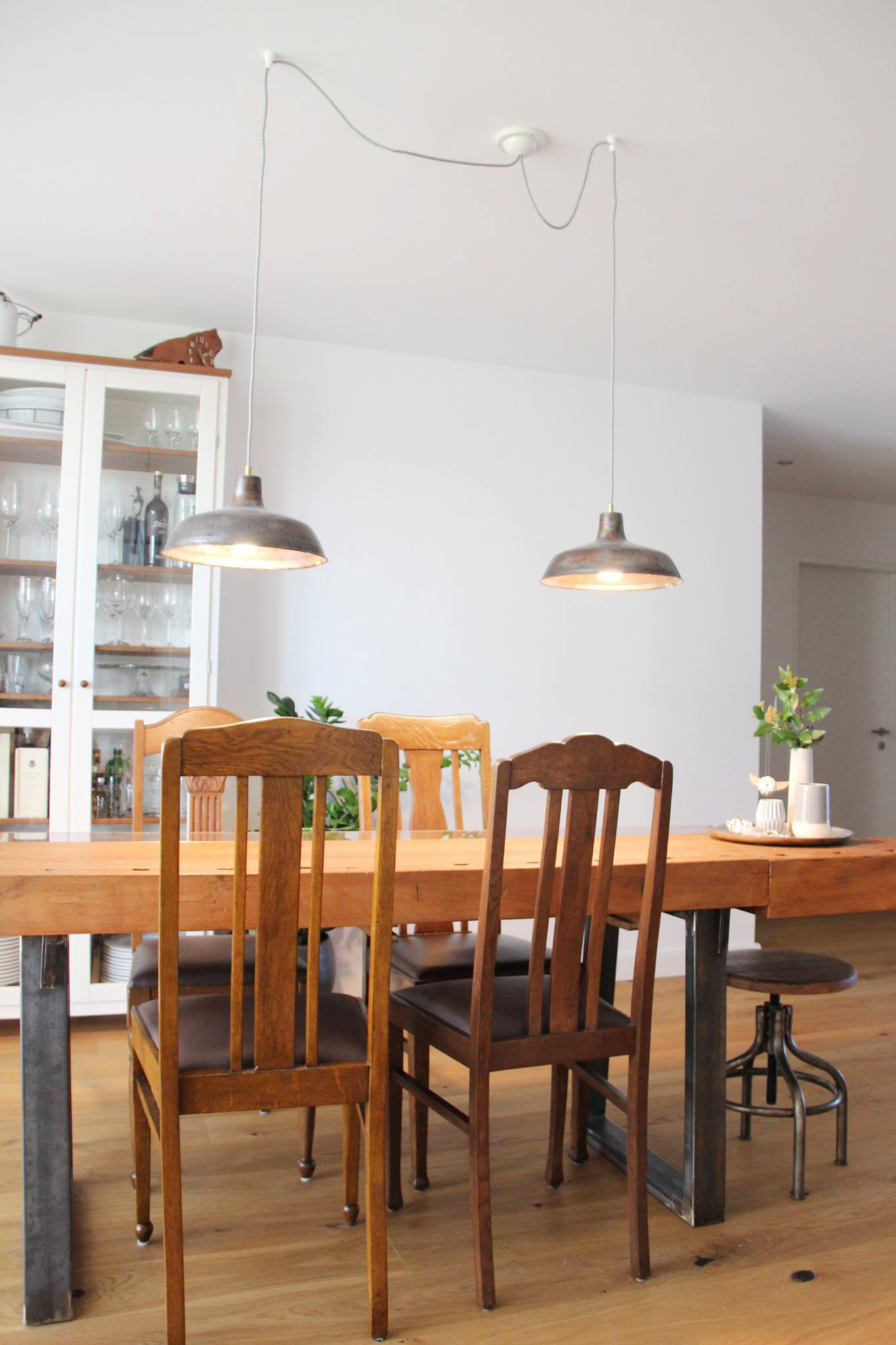 Ein Esszimmer, in der Mittevsteht ein Holztisch aus einer alten Werkbank mit vier antiken Holzstühlen darum. Über dem Tisch hängen zwei Fabrikleuchten aus Metall, die an der Decke an einem Anschluss angeschlossen sind